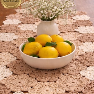 Puede incluir: Camino de mesa de ganchillo en tonos beige y marrón, con un diseño floral. Un cuenco de cerámica blanca contiene varios limones amarillos. Un jarrón blanco con flores blancas también está sobre la mesa. El texto "EASY/PDF TABLE RUNNER" está en la esquina superior izquierda.
