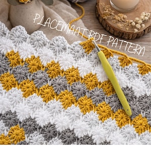 Può includere: Un tessuto parzialmente lavorato all'uncinetto con un motivo ripetitivo a forma di stella in filato giallo senape, bianco e grigio. Un uncinetto verde chiaro è inserito nel lavoro. Il testo PLACEMAAT PDF PATTERN è sovrapposto all'immagine.
