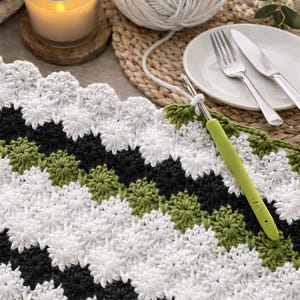 May include: A close-up of a handmade crochet project in progress. The piece features a repeating pattern of white, black, and green floral motifs. A green crochet hook is in use. A lit candle, a ball of yarn, and a plate with silverware are in the background.