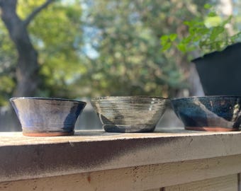 May include: Three handmade ceramic bowls with a rustic aesthetic. The bowls are in shades of blue, gray, and green, with visible texture and a natural, earthy feel. Each bowl has a unique shape and color variation, perfect for serving or display.