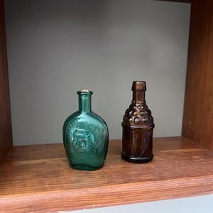 May include: Two small glass bottles, one emerald green and one amber brown. The green bottle has a flattened, rounded shape with embossed details, while the brown bottle features a textured, grape-like pattern on the upper body and a narrow neck.