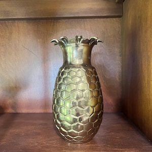 May include: A metallic gold-toned brass vase shaped like a pineapple. The vessel features a textured, repeating hexagonal pattern across its body and a flared, scalloped rim at the top, mimicking the leaves of a pineapple fruit structure.