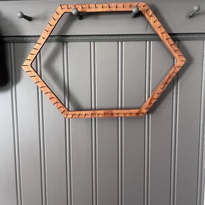 May include: A hexagonal wooden weaving loom frame featuring a series of evenly spaced metal pins protruding from the outer edges. The frame is constructed from light brown wood with a visible grain, designed for yarn or thread craft projects.