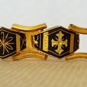 May include: A gold-tone bracelet with black enamel links. Each link features a different geometric design, including a cross, a star, and a diamond shape.