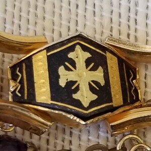 May include: A gold-toned metal bracelet with a black enamel center piece featuring a gold cross design. The center piece has a textured border with gold accents.