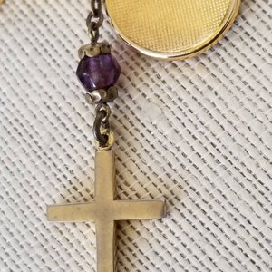 May include: A gold cross pendant with a purple glass bead charm hanging from a gold chain.