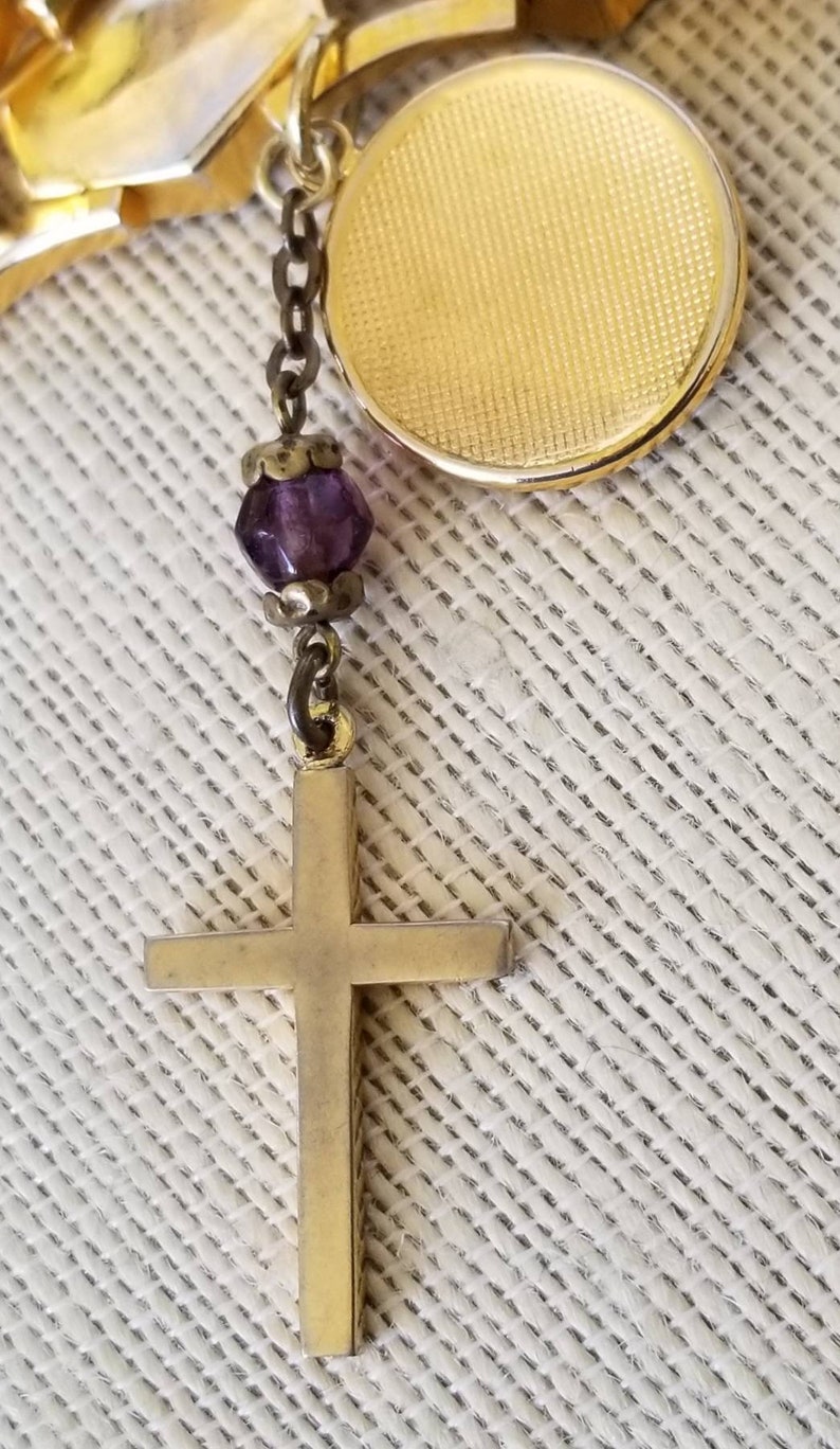 May include: A gold cross pendant with a purple glass bead charm hanging from a gold chain.
