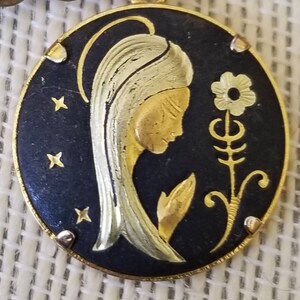 May include: A gold-toned pendant featuring a black enamel oval with a gold silhouette of a woman with a flower. The woman's hair is white and gold, and the flower is stylized with a gold stem and leaves.