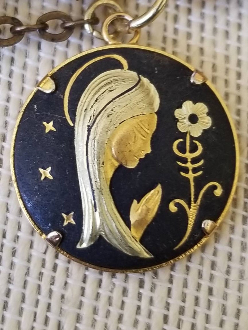 May include: A gold-toned pendant featuring a black enamel oval with a gold silhouette of a woman with a flower. The woman's hair is white and gold, and the flower is stylized with a gold stem and leaves.