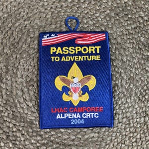 May include: A rectangular blue embroidered patch featuring a red and white striped flag graphic at the top. The text reads PASSPORT TO ADVENTURE, LHAC CAMPOREE, ALPENA CRTC, 2004. A yellow fleur-de-lis emblem with a central eagle is centered on the patch.