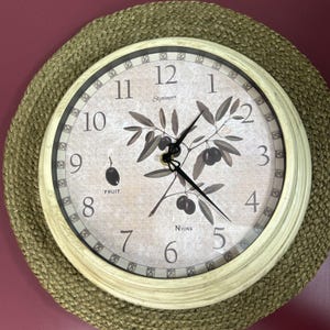 May include: Cream-colored wall clock with black hands and numbers, featuring an olive branch design. The clock face has the words "Skytimer" and "Nyons" printed on it. The clock is set on a woven, circular placemat.