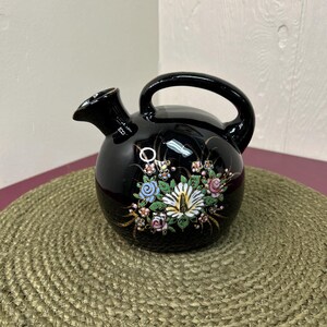 May include: A black ceramic pitcher with a round body, a handle, and a spout. The pitcher is decorated with a floral design in shades of pink, blue, and white. The pitcher is sitting on a woven mat.