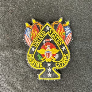 Può includere: Toppa ricamata nera e gialla a forma di picche, con la scritta "United States Marine Corps". La toppa include un'aquila, bandiere e stelle, con un bordo giallo.
