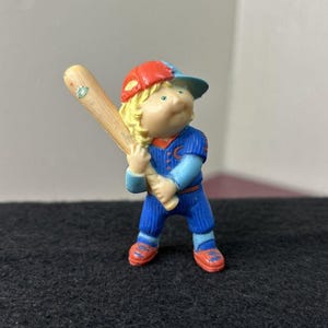 May include: A figurine of a child in a baseball uniform, holding a wooden bat. The figure wears a blue uniform with red accents, a red and blue cap, and red shoes. The figure has blonde hair and is in a batting stance.