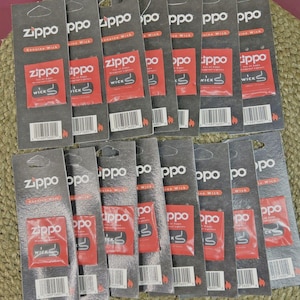 May include: A collection of Zippo lighter wick replacement packs. Each pack features the Zippo logo and the word "wick" on a red background. The packs are gray and arranged on a woven surface.