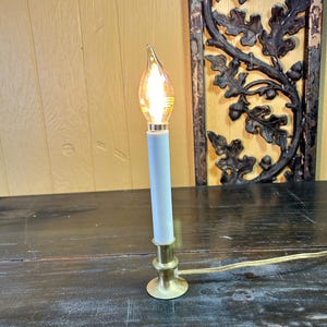 May include: A small electric candle lamp with a light blue body and a brass base. The bulb is shaped like a flame and emits a warm, yellow light. A gold-colored cord extends from the base.