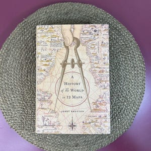 May include: A hardcover book titled "A History of the World in 12 Maps" with a vintage map design. The cover features a compass illustration and the author's name, Jerry Brotton. The book is set on a woven placemat.