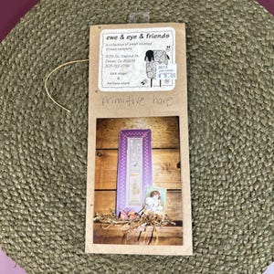 May include: A tan paper packaging sleeve labeled ewe & eye & friends with a printed image of a purple vertical sampler featuring a hare illustration and text. The label includes the address 1039 So. Gaylord St., Denver, Co, 80209 and phone number.