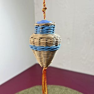 May include: A hanging woven ornament with a light blue top and accents. The main body is a natural tan color with blue bands. A tassel of red and yellow threads hangs from the bottom, and a twisted cord suspends the ornament.