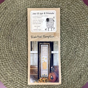 May include: A tan paper craft pattern packet titled ewe & eye & friends, a collection of small counted thread samplers, featuring a Primitive Pumpkin design. The image shows a vertical sampler with an alphabet, yellow stars, and a pumpkin.