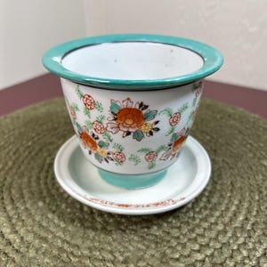 May include: A white ceramic planter with a turquoise rim and base, adorned with orange and yellow floral patterns. The planter sits on a matching saucer, creating a cohesive and decorative set. The design is detailed and colorful.