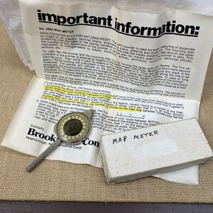 May include: A silver map measuring tool with a dial and a black and white instruction sheet. The instruction sheet reads "Important Information: No. 2962 Map Meter Inch Counter with Instant Zero Reset".