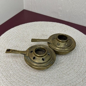 May include: Two small brass incense burners with handles. The burners have a round shape with a small hole in the center and several smaller holes around the edge.