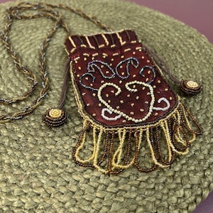 May include: A small, rectangular, deep red pouch necklace featuring intricate beadwork in gold, silver, and dark tones. The pouch has a drawstring closure with beaded ends, a decorative beaded fringe at the bottom, and a long, twisted beaded cord.
