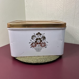 May include: A vintage white metal bread box with a gold-colored lid. The front of the box features a floral design in brown and red tones. The box is rectangular and sits on a green woven mat.