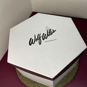 May include: A white, hexagonal box with the text "Wolf Wile's LEXINGTON, KY." in black script. The box has a dark brown trim and sits on a green woven mat, against a burgundy surface.