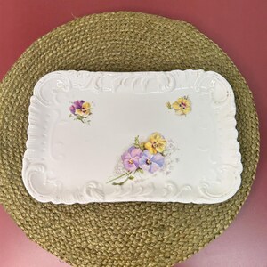 May include: A white rectangular serving tray with a floral design. The tray has a decorative, scalloped edge and is adorned with painted purple, yellow, and white pansies. The tray is placed on a round, woven, tan placemat.