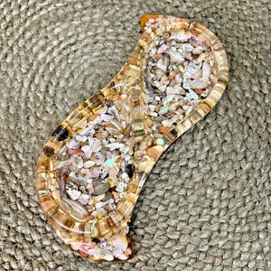 May include: A decorative, resin tray with a unique design. The tray is filled with small, colorful stones and shells. The edges of the tray have a striped pattern in shades of brown and black. The tray is a light amber color.
