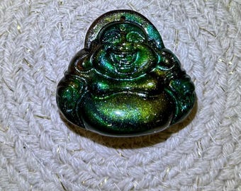 May include: A metallic green and gold Buddha pendant with a smiling face. The pendant has a loop at the top for hanging and is set against a textured, off-white background. The Buddha figure is detailed with rounded features.