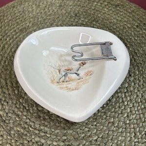 May include: Cream-colored, heart-shaped ceramic dish with a hunting dog illustration. A silver-colored metal clip is attached to the dish. The dog is white with brown spots, set against a backdrop of tall grass.
