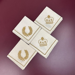 May include: Four white linen napkins with gold embroidered designs. Two napkins feature a crown with three stars and two napkins feature a laurel wreath.