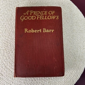 May include: A red hardcover book with gold lettering. The title is "A Prince of Good Fellows" by Robert Barr.
