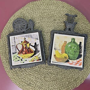 May include: Two vintage trivets with black metal frames and ceramic tile inserts. One features a teapot, the other a green bottle and pears. Both are on a woven mat, showcasing a rustic, farmhouse aesthetic.