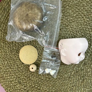 May include: A collection of doll-making supplies, including a doll head, wig, and various small parts. The doll head is pale pink, and the wig is a light brown color. Other parts include a small wooden bead and a round, yellow fabric piece.