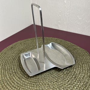 May include: A stainless steel spoon rest featuring a vertical handle and two distinct recessed compartments for holding kitchen utensils. The base is rectangular with rounded edges, and the upright loop is made of thin, polished metal wire.