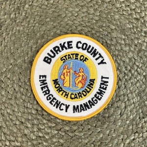 May include: A circular patch with the text "BURKE COUNTY EMERGENCY MANAGEMENT" in black lettering on a white background. The center features a gold and blue emblem with the words "STATE OF NORTH CAROLINA". The patch has a yellow border.