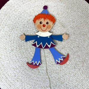 May include: A wooden pull-string clown toy with a blue outfit, red hair, and a smiling face. The clown has a white collar, purple accents, and red shoes. The toy is set against a textured, off-white background.