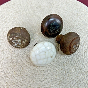 May include: Four antique door knobs in various styles. Two are aged metal with floral designs, one is a dark brown, and the fourth is a white, cracked ceramic. The knobs are arranged on a textured, off-white surface.