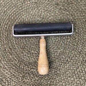 May include: A black rubber brayer with a silver metal frame and a light wood handle. The brayer is designed for printmaking and crafting. The handle is a light tan color.