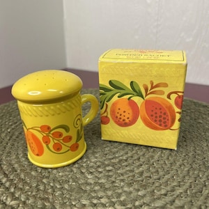 May include: A yellow ceramic salt shaker with a handle and a matching box. Both items feature a fruit design in orange and green. The salt shaker has a perforated top. The box reads "POWDER SACHET".