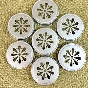 May include: Seven silver-colored, round jar lids with a decorative floral pattern cut into the center. The lids are arranged in a circular pattern on a woven, tan surface. The lids have a slightly distressed appearance.
