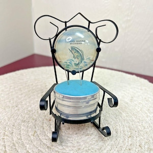 May include: A vintage black metal chair-shaped pin cushion. The chair has a round glass back with a picture of a fish jumping out of the water and the words "Grand Rapids Michigan". The cushion is blue and the chair is sitting on a white woven mat.