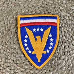 May include: A shield-shaped patch with a blue background, featuring a gold eagle and white stars. The top border has red and white stripes, and the patch is outlined in gold. This patch is likely a military or patriotic emblem.