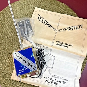 May include: A vintage Videoman Teleporter kit with a telescoping antenna, AC-DC adapter, and instructions. The device has a blue and silver face with the word "VIDEOMAN" and power settings. The kit includes a black cable and is on a tan paper.