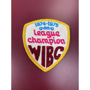 May include: White felt patch with yellow stitching, featuring the text "1974-1975" in blue, a swirl design, "league champion" in pink and red, and "WIBC" in maroon.