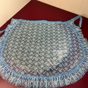 May include: A vintage blue apron with a floral pattern and ruffled trim. The apron is made of a clear, waterproof material with white flower designs. The edges are trimmed with a blue and white lace-like border, and it has blue ties.